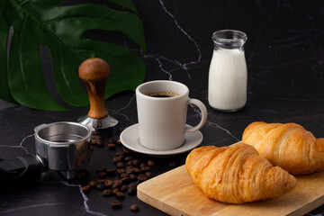 A white coffee cup was placed next to a milk bottle and a croissant on a cutting board, coffee beans and grinders on a marble floor.
