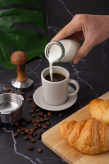 Fototapeta na wymiar The hand was holding a milk bottle pouring it into a white coffee cup, croissant on a cutting board, coffee beans and a coffee grinder on a black marble floor.