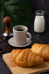 A white coffee cup was placed next to a milk bottle and a croissant on a cutting board, coffee beans and grinders on a marble floor.