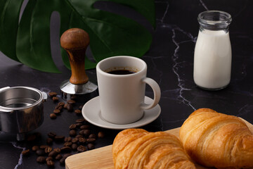 A white coffee cup was placed next to a milk bottle and a croissant on a cutting board, coffee beans and grinders on a marble floor.