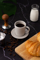 A white coffee cup was placed next to a milk bottle and a croissant on a cutting board, coffee beans and grinders on a marble floor.
