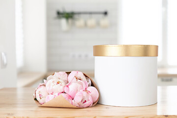 Gold gift box mockup on a wooden table in the kitchen. Glass vase with a bouquet of pink peonies. Scandinavian style interior.
