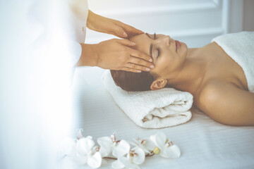 Obraz na płótnie Canvas Beautiful brunette woman enjoying facial massage with closed eyes in sunny spa center. Relaxing treatment and cosmetic medicine concepts