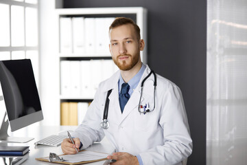 Friendly red-bearded doctor sitting and writing at clipboard in clinic. Perfect medical service in hospital. Medicine, healthcare