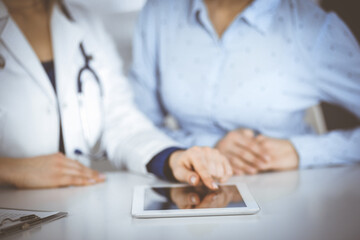 Unknown woman-doctor is showing to her patient a description of medication, while sitting together at the desk in the cabinet in a clinic. Female physician is using a computer tablet and a stethoscope