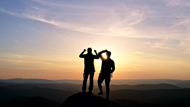 Silhouettes of a happy young couple on top of a mountain triumphantly raising their hands in the air