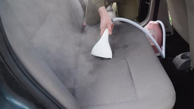 Auto detailing of an old used car. The interior of the car is cleaned with a steamer. A man in work clothes cleans the rear seats of the car from dust and dirt.