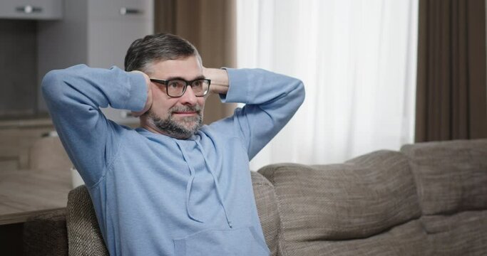 A man with gray hair in glasses closes his ears with his hands reacts negatively can no longer listen aggressively shakes his head sitting on the couch at home