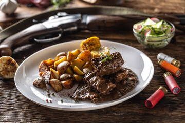 Wild game goulash stew served with roasted potatoes and seasoned with four colour pepper corn. Whole food is surrounded by hunting shotgun and gun bullets.