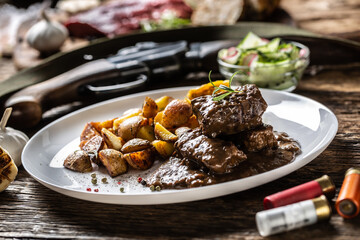 Wild game goulash stew served with roasted potatoes and seasoned with four colour pepper corn. Whole food is surrounded by hunting shotgun and gun bullets.