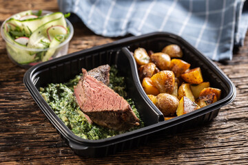 Delicious red meat sous vide steak packed in portable take away plastic box and served with crunchy roasted potatoes and savory spinach dip and fresh vegetable salad