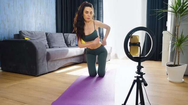 Sporty woman doing cat cow exercise, arching back, recording video with smartphone on tripod. Fitness blogger at online training during lockdown. Concept of blogging