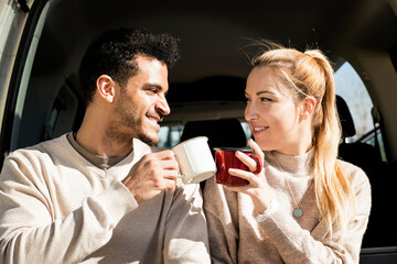 Happy young couple having a coffee break during road trip in countryside. Man and woman sitting in car trunk and having coffee.