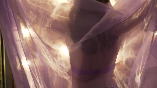 Young slender woman dancing slow dance with her back in purple dress with a transparent shawl in a room in the evening