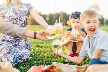 Family with two brothers, mother and father summer picnic in nature. Little children with parents hold glasses with cold lemonade in their hands. Kids boys drinking a refreshing drink from the straw.  - obrazy, fototapety, plakaty