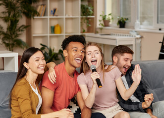 Hang out with friends. Cheerful young multicultural friends playing karaoke at home, singing with microphone, having great time together while sitting on the couch in the living room