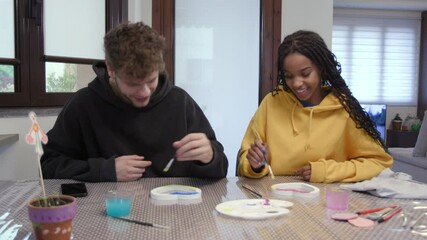 Man and woman smiling while painting wooden hearts at home. Domestic life for mixed couple doing leisure activity with paint for hobby. Happy people laughing on Valentine's Day