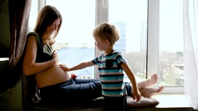 Pregnancy woman sitting near window at home, little child boy older brother touching and kissing pregnant mother belly. Authentic family moments of expecting baby.