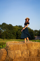 Naklejka na ściany i meble A young beautiful woman in a straw hat stands on a large pile of straw bales. Freedom, wind, setting sun.