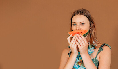 smiling caucasian girl holding slice of watermelon in front her mouth on brown background with copyspace.