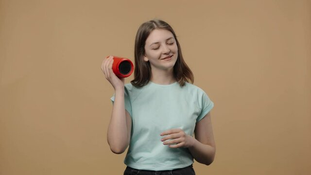 Portrait of charming beauty model listening to music with bluetooth portable speaker and dancing. Young girl in white t-shirt posing on brown studio background. Close up. Slow motion ready 59.94fps.