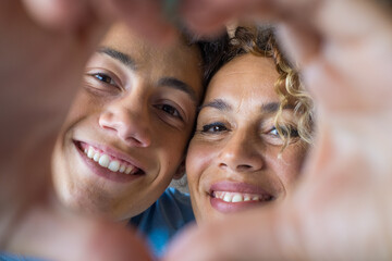 Portrait of couple of mom and son together a home making a heart love shape with their fingers and hands with the faces at the background. Affectionate teenager and woman enjoying
