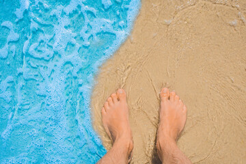 The young man's legs stand on the beach sand and are washed by blue sea water, a view from above