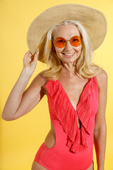 Fototapeta premium Studio shot of happy blonde woman in sunglasses, red swimsuit and straw hat smiling at camera, adjusting her hat while posing, standing isolated on yellow background