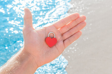 In the hand of a man on the palm of a small red heart close-up in the form of a lock. A sign or symbol of love against the sea coast