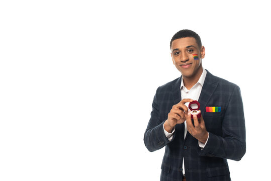smiling african american man with lgbt flag painted on face holding wedding ring isolated on white