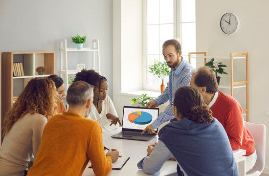 Diverse Team Meeting Around Office Table. Business Group Leader Making Presentation, Showing Sales Statistics Or Feedback Report Data And Explaining What Circle Pie Charts On Laptop Screen Stand For