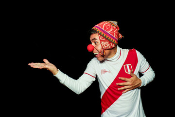 A clown smiles happily with one hand extended and the other on his chest, wearing an Andean hat and a Peruvian sports shirt.