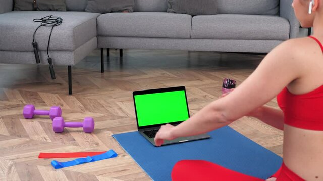 Green screen mock up chroma key monitor laptop concept: Fit woman in sportswear sits on yoga mat looks computer display, tells fitness trainer online video call webcam, distance sports lesson course