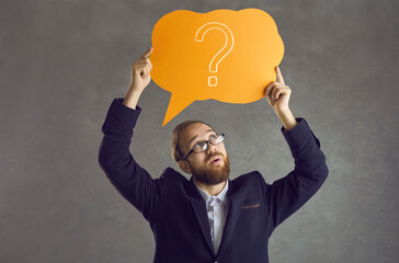 Serious sceptical smart young business man in glasses with dubious judging evaluating face expression holding orange speech bubble with question mark and thinking isolated on grey background