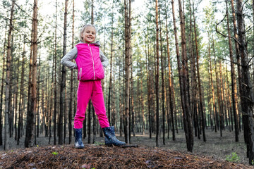 Cute pretty blond caucasian happy child funny girl in bright pink suit enjoy walking in wet coniferous pine forest on sunny morning day. Little female kid portrait hiking nature trip outdoors