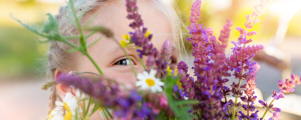 Close-up portrait of little cute caucasian blond kid girl peeking and hiding holding bouquet of salvia sage wild flowers walking at grass meadow outdoors summer day. Young female person wildflowers