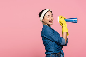 Smiling housewife in rubber gloves holding detergent and winking at camera isolated on pink