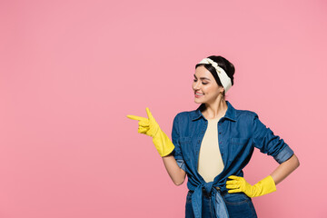Cheerful woman in rubber gloves pointing with finger isolated on pink