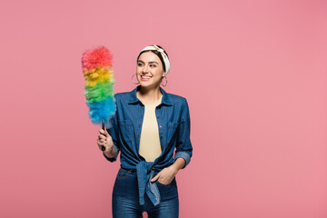 Smiling housewife looking at dust brush isolated on pink