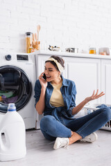 Positive housewife talking on smartphone near detergent and washing machine