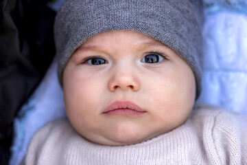 Portrait of serious baby in grey hat. Sweet little infant looks to camera. Kid with brown eyes and long lashes