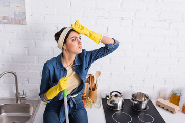 Exhausted housewife in rubber gloves holding mop on kitchen worktop