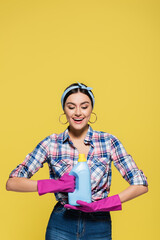 Smiling woman in rubber gloves looking at bottle of detergent isolated on yellow