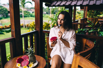 Dreamy woman enjoying ripe pitaya on wooden terrace in morning