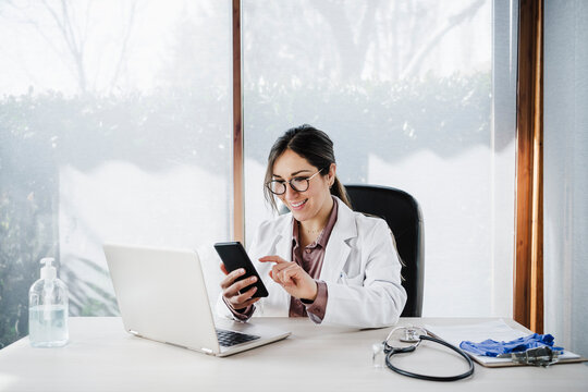 Smiling female doctor using mobile phone while sitting at desk in hospital - Powered by Adobe