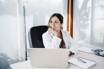 Smiling female medical professional with hand on chin day dreaming while sitting at desk