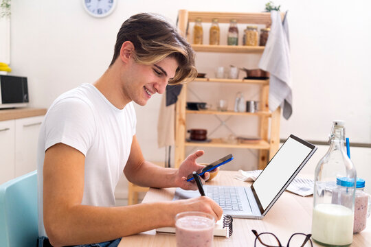 Happy man writing on note pad while siting with laptop and mobile phone in kitchen