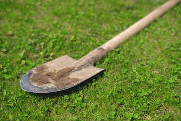 A side view of a dirty shovel lying on the grass, illuminated by the bright rays of the sun and casting a shadow.