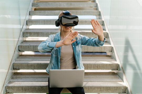 Woman with laptop gesturing while watching video through virtual reality simulator on steps