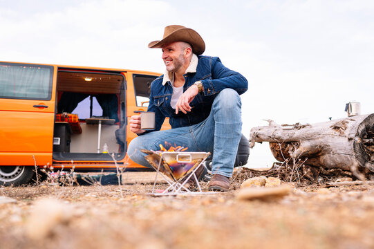 Smiling mature man in hat crouching while having coffee by wood burning stove during camp